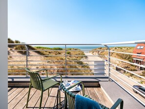 Un balcon avec vue sur la plage et les dunes, comprenant deux chaises et une table avec un journal.