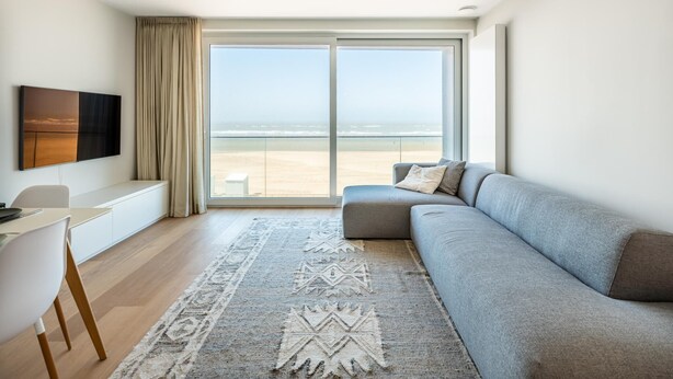 A modern living room with a large window, a grey sofa, a white desk, and a patterned rug. - Newly built flat with sea view (Koksijde)