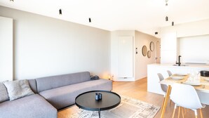 A modern living room with a grey sofa, a round coffee table, and a dining area with white chairs and a table set for two. - Newly built flat with sea view (Koksijde)