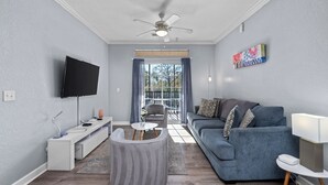 A modern living room with a grey sofa, a white entertainment unit, a ceiling fan, and a large window with curtains.