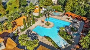 An aerial view of a resort with a central swimming pool, surrounded by palm trees and outdoor seating areas.