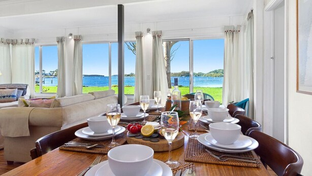 A dining area with a table set for a meal, large windows with a view of the sea, and a sofa in the background. - Noddy`s Charming Waterfront Cottage (Greenwell Point)