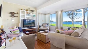 A spacious living room with a television, seating area, and a view of the lake. - Noddy`s Charming Waterfront Cottage (Greenwell Point)