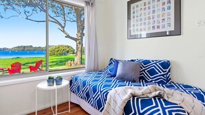A bedroom with a large window, a bed with blue and white bedding, a small table with two potted plants, and a framed photo collage on the wall. - Noddy`s Charming Waterfront Cottage (Greenwell Point)