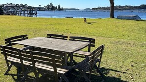 A picnic table with benches in a grassy area with a view of the water and a tree. - Noddy`s Charming Waterfront Cottage (Greenwell Point)