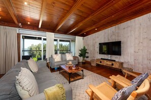 Ein geräumiges Wohnzimmer mit einer großen, grauen Couch, einem Couchtisch und Holzstühlen. An der Wand ist ein Flachbildfernseher befestigt, und ein großes Fenster bietet einen Blick ins Freie.