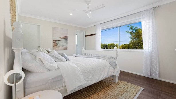 A bedroom with a bed, a bedside table, a window with curtains, a ceiling fan, and a painting on the wall. - Heated Pool, Fireplace, Games Room & Pet Friendly! (Sanctuary Point)