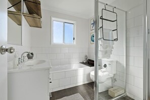 A modern bathroom with a white sink, bathtub, and toilet. There is a window above the sink and a shower area with a glass door. - Heated Pool, Fireplace, Games Room & Pet Friendly! (Sanctuary Point)