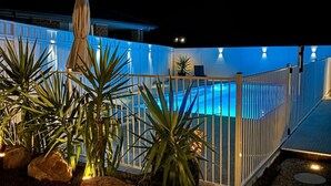 A nighttime view of a pool area with a white fence, potted plants, and stepping stones leading to the pool. - Heated Pool, Fireplace, Games Room & Pet Friendly! (Sanctuary Point)