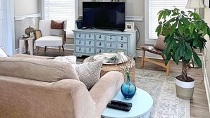 Una sala de estar con un ventilador de techo, un televisor sobre un mueble azul claro, un espejo grande y una planta en maceta.