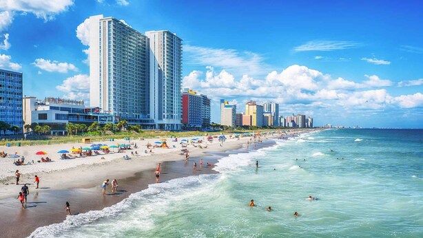 Una playa con una fila de hoteles altos, cielos despejados y gente disfrutando.