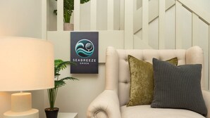Una sala de estar con un sillón color beige, una lámpara y un logotipo enmarcado en la pared.