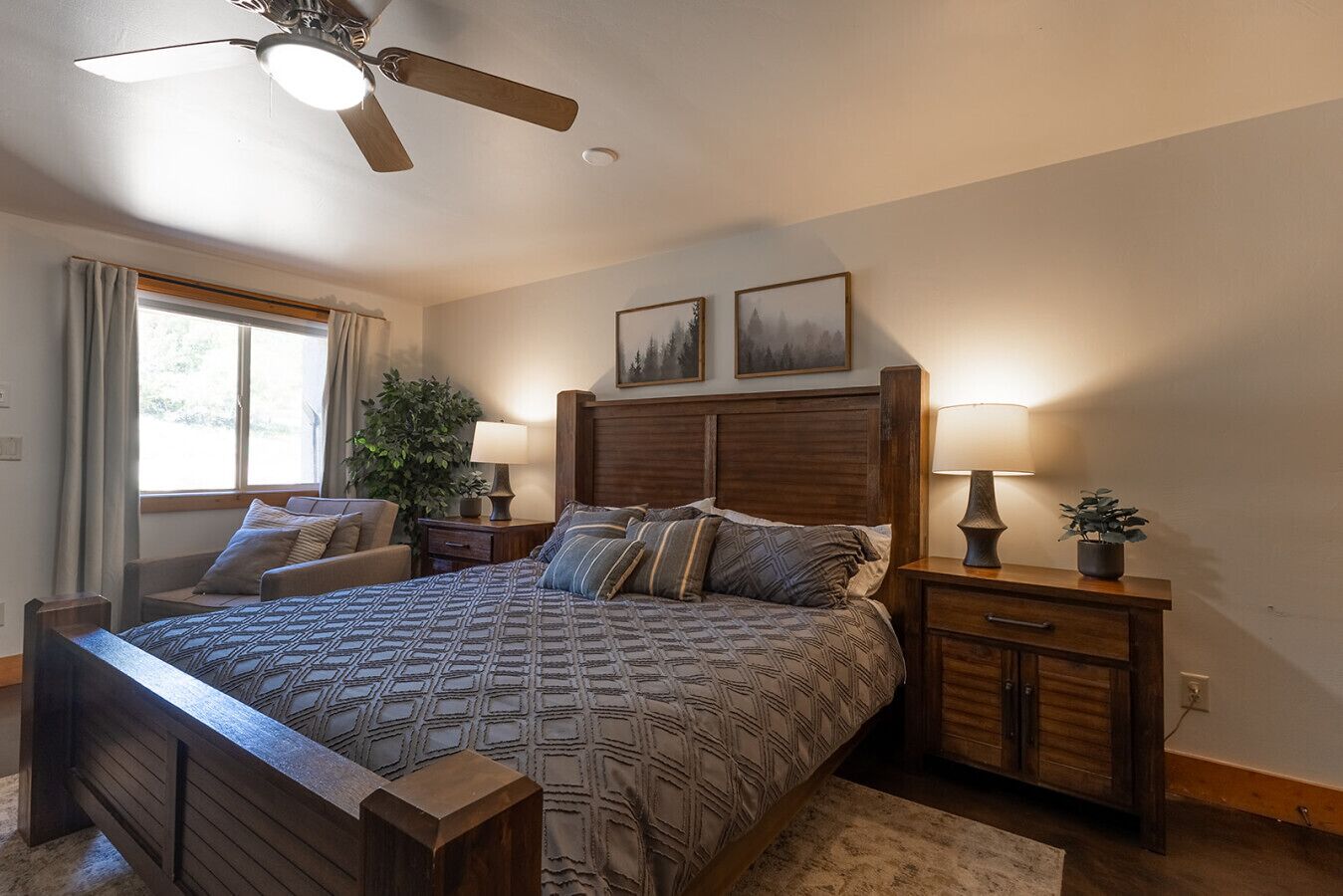 A bedroom with a large bed, two bedside tables, a ceiling fan, a window with curtains, and two framed pictures on the wall.