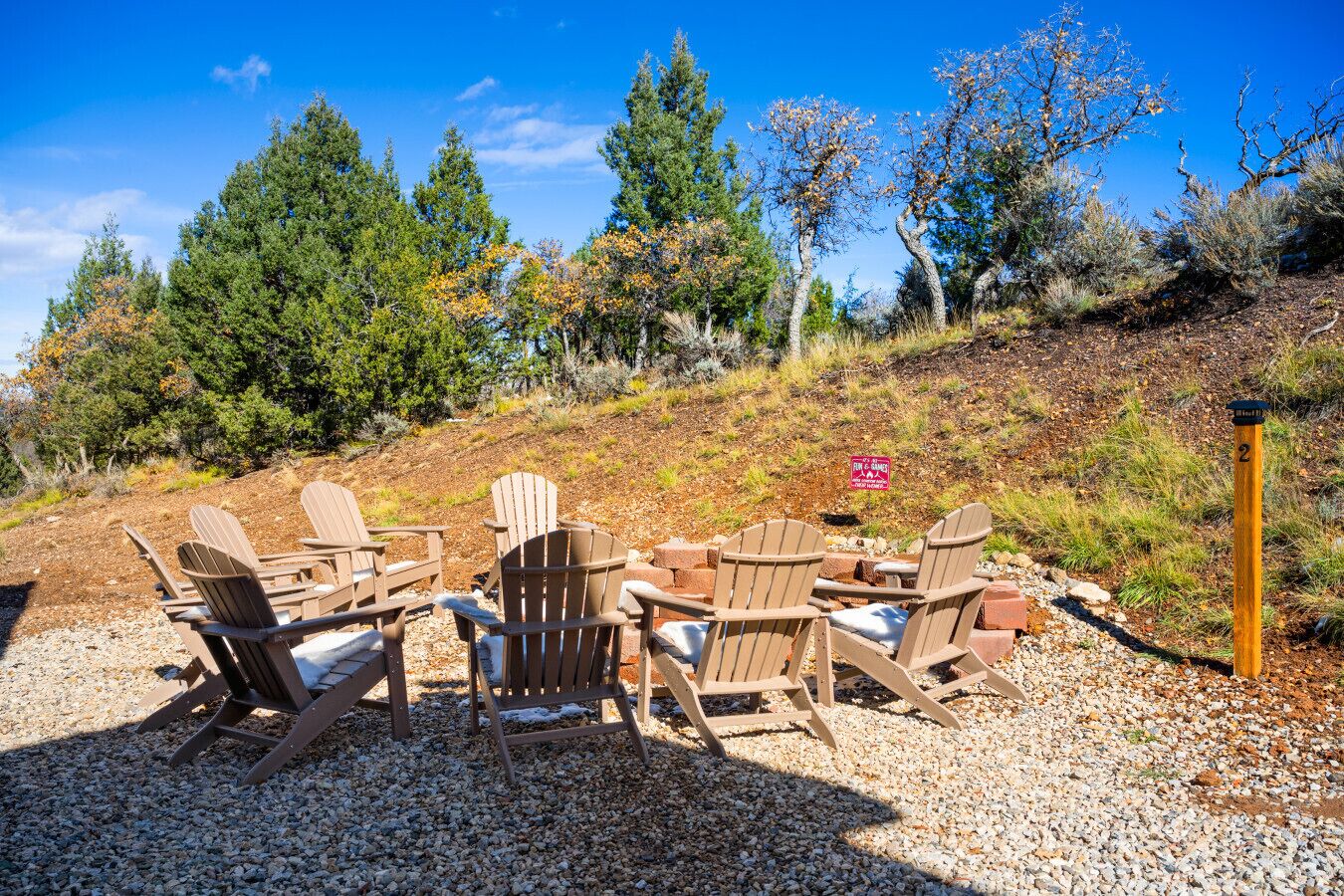 Eine Sitzecke mit Adirondack-Stühlen und einer Feuerstelle, umgeben von Bäumen und blauem Himmel.