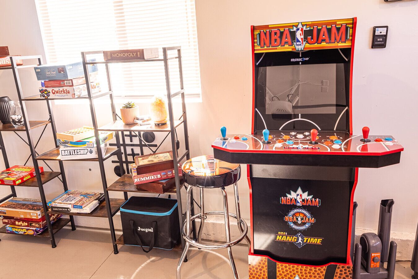 Ein Zimmer mit einem Monopoly-Spiel auf einem Regal, einer NBA JAM Spieluhr und einer Tasche mit dem Aufdruck 'Jugol'.