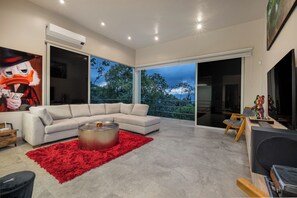 A modern living room with a white sectional sofa, a red shag rug, and a large window offering a view of trees and a cloudy sky.