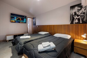 A hotel room with two beds, a wooden headboard, a nightstand with a lamp, a painting of a woman on the wall, and a cityscape on the TV.