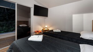 Et moderne hotelværelse med to senge, et fjernsyn ophængt på væggen, et lille bord med en lampe og en skydedør, der fører ud til en balkon med udsigt over grønne områder.