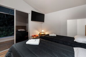 Ein modernes Hotelzimmer mit zwei Betten, einem an der Wand befestigten Fernseher, einem kleinen Tisch mit Lampe und einer Schiebetür zu einem Balkon mit Blick auf Grünflächen.