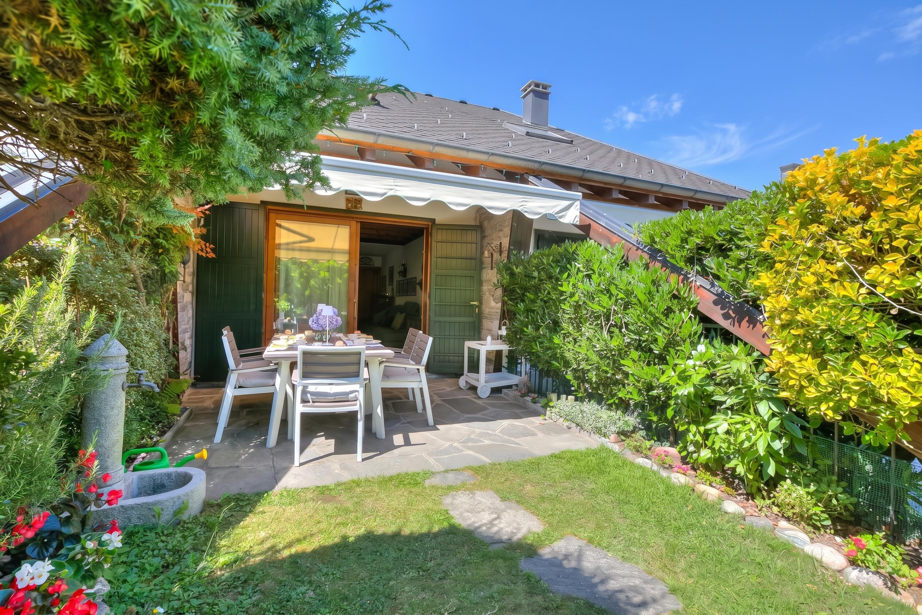 A patio with a table and chairs, surrounded by a garden with various plants and a stone pathway.