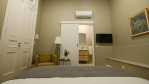 A hotel room with a bed, a chair, a small table with a plant, a bathroom with a mirror, and a TV mounted on the wall.