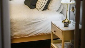 En pænt redt seng med hvide sengetæpper og stribede puder, en sengetøjslampe og et lille natbord med lampe og en vase med blomster.