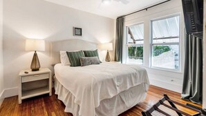Un dormitorio con cama, dos mesitas de noche, ventilador de techo, ventana con cortinas y vista a zonas verdes.