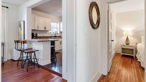 Cocina compacta con armarios blancos, comedor con dos sillas, una pequeña isla y un pasillo que lleva a un dormitorio con mesita de noche y lámpara.