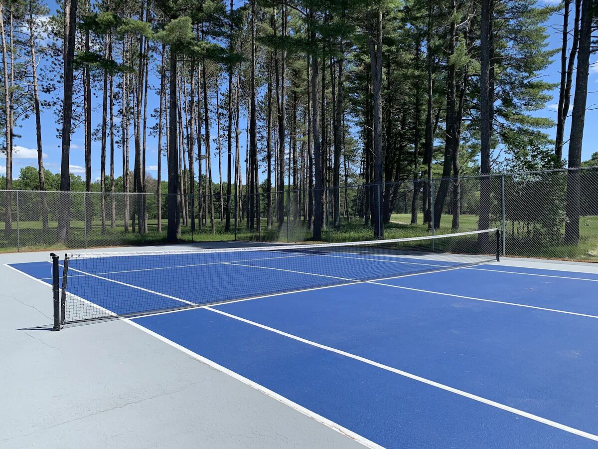 An outdoor tennis court with a blue surface and white boundary lines, surrounded by a chain-link fence and tall trees.