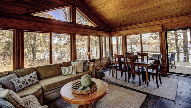 Een gezellig interieur van een chalet met houten plafond, grote ramen, een bank, een eettafel en stoelen.