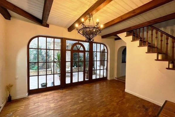 A spacious room with a wooden floor, large arched windows, a chandelier, and a staircase.