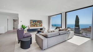 Un salon moderne avec un grand canapé gris, une petite table d’appoint, un téléviseur et une grande fenêtre donnant sur la mer.