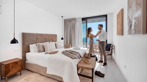 Une chambre moderne avec un grand lit, une petite table de chevet et une vue sur la mer, visible à travers une porte coulissante.