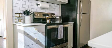 A compact kitchen with white cabinets, a black oven, a stainless steel refrigerator, and a tiled backsplash.