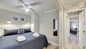 Een netjes opgemaakt bed met een donkerblauwe dekbedsprei, twee kussens en opgevouwen witte handdoeken. Boven het bed is een plafondventilator zichtbaar.