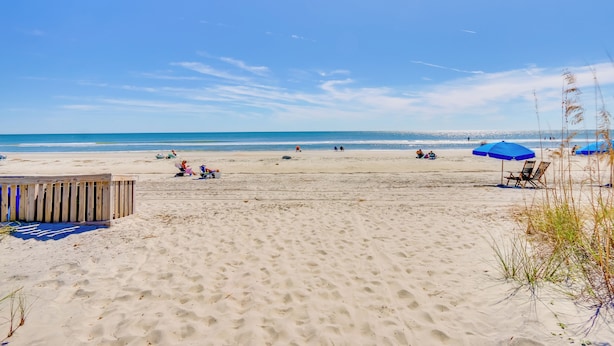 Ein Sandstrand mit einer Holzkonstruktion, blauen Sonnenschirmen und blauem Himmel.