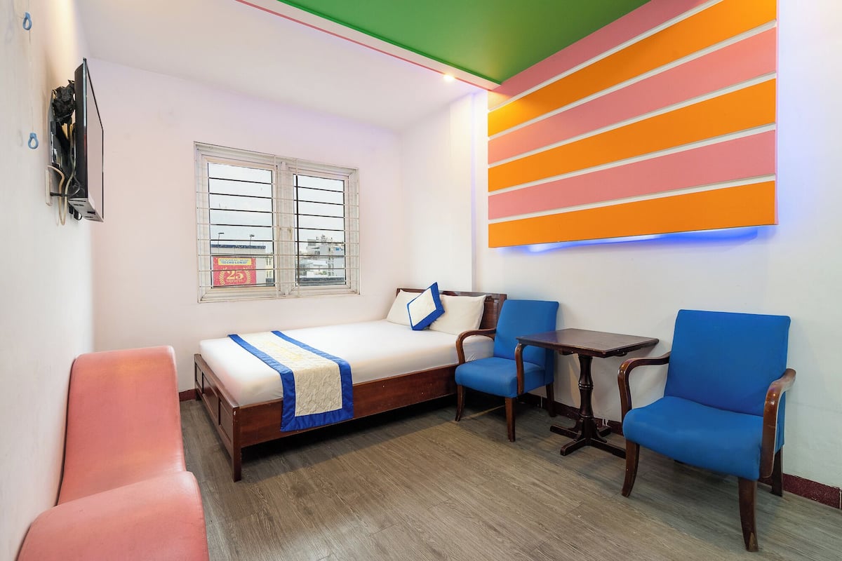 A room with a bed, two chairs, a small table, a window with blinds, and a colorful wall decoration.