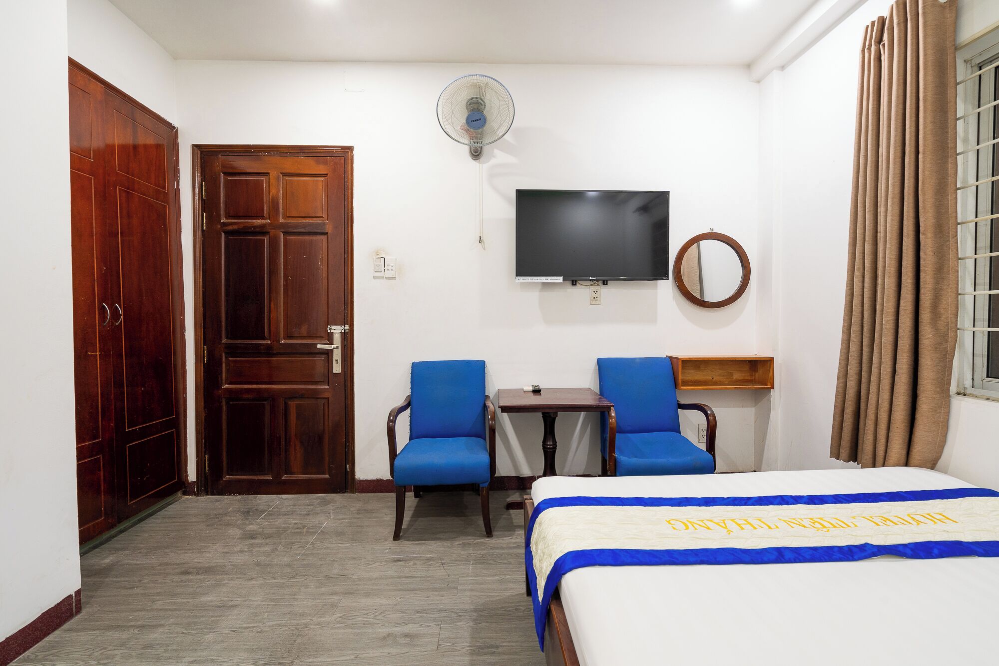 Ein Hotelzimmer mit einem Bett, zwei blauen Stühlen, einem kleinen Tisch, einem Fernseher und einem Ventilator.
