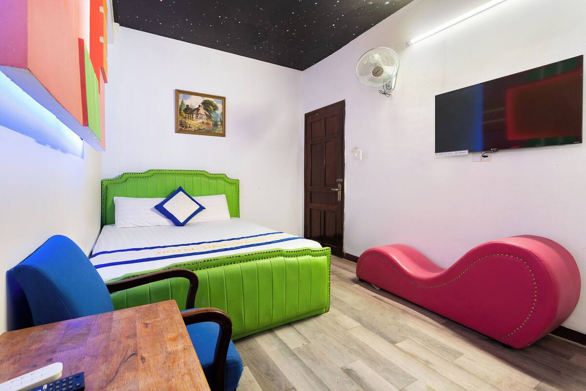 A bedroom with a green upholstered bed, a blue chair, a wooden table, a pink sofa, a wall-mounted TV, and a ceiling fan.