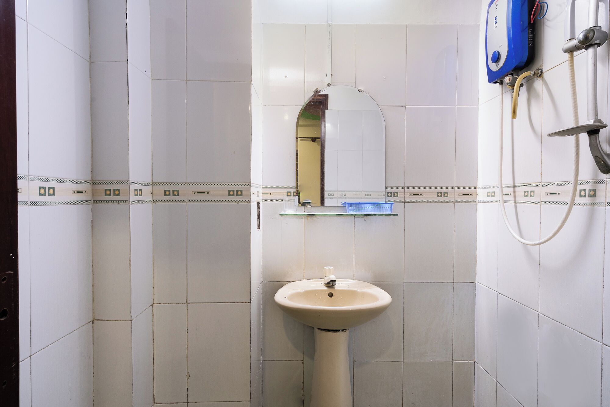 A bathroom with white tiles, a pedestal sink, a mirror, and a shower control panel.