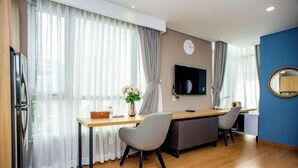 A modern hotel room with a wooden desk, two chairs, a flat-screen TV, and a large window with sheer curtains. - Lancaster Ho Chi Minh Serviced Apartment Official (Ho Chi Minh City)