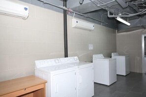 A laundry room with three white washers and dryers, a wooden bench, and exposed ductwork. - Lancaster Ho Chi Minh Serviced Apartment Official (Ho Chi Minh City)
