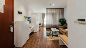 A modern living room with a sofa, coffee table, and a plant. There is a kitchen area with a refrigerator and a counter. The room has wooden flooring and a striped curtain in the background. - Lancaster Ho Chi Minh Serviced Apartment Official (Ho Chi Minh City)