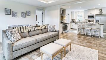 A modern living room with a grey sofa, a coffee table, and a kitchen area with white cabinets and stainless steel appliances.