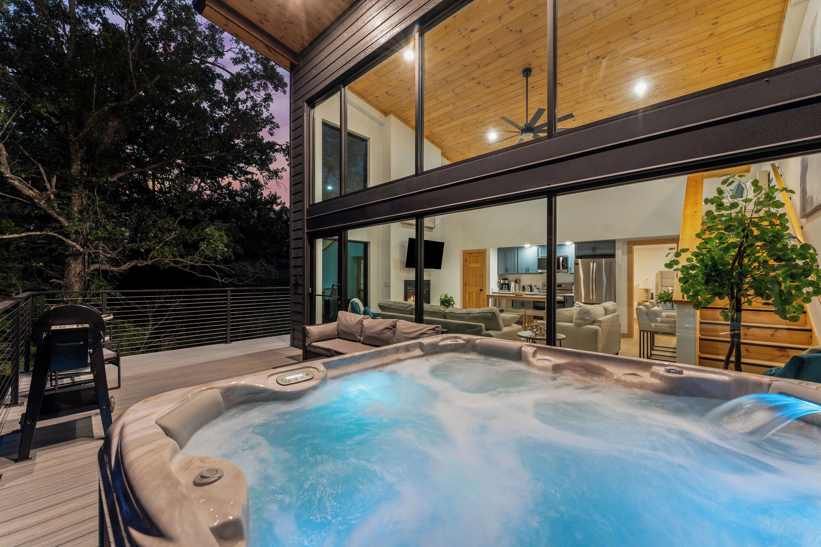 A modern outdoor hot tub area with a wooden deck, a seating area with sofas, and a view of the interior of a house through large windows.