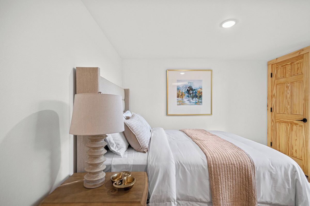 A bedroom with a bed, a bedside table with a lamp, a framed picture on the wall, and a wooden wardrobe.