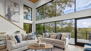A modern living room with two sofas, a coffee table, and large windows offering a view of trees and a balcony.