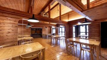 A wooden interior with multiple dining tables and chairs, a kitchen area, and large windows allowing natural light.