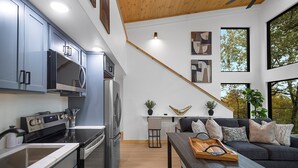 Una cocina moderna con electrodomésticos de acero inoxidable, un comedor con un sofá oscuro y una mesa de madera, y ventanas amplias que ofrecen vistas a árboles.