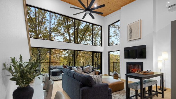 Una sala de estar moderna con un sofá grande, una mesa de comedor con taburetes tipo bar, una chimenea y ventanas amplias con vista a árboles.